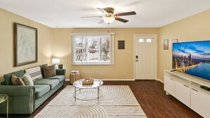 A living room with a green sofa, a glass coffee table, a ceiling fan, a large TV, and a window with blinds. - Sailor`s Escape with 3 bedrooms Near Naval Base! (Waukegan)