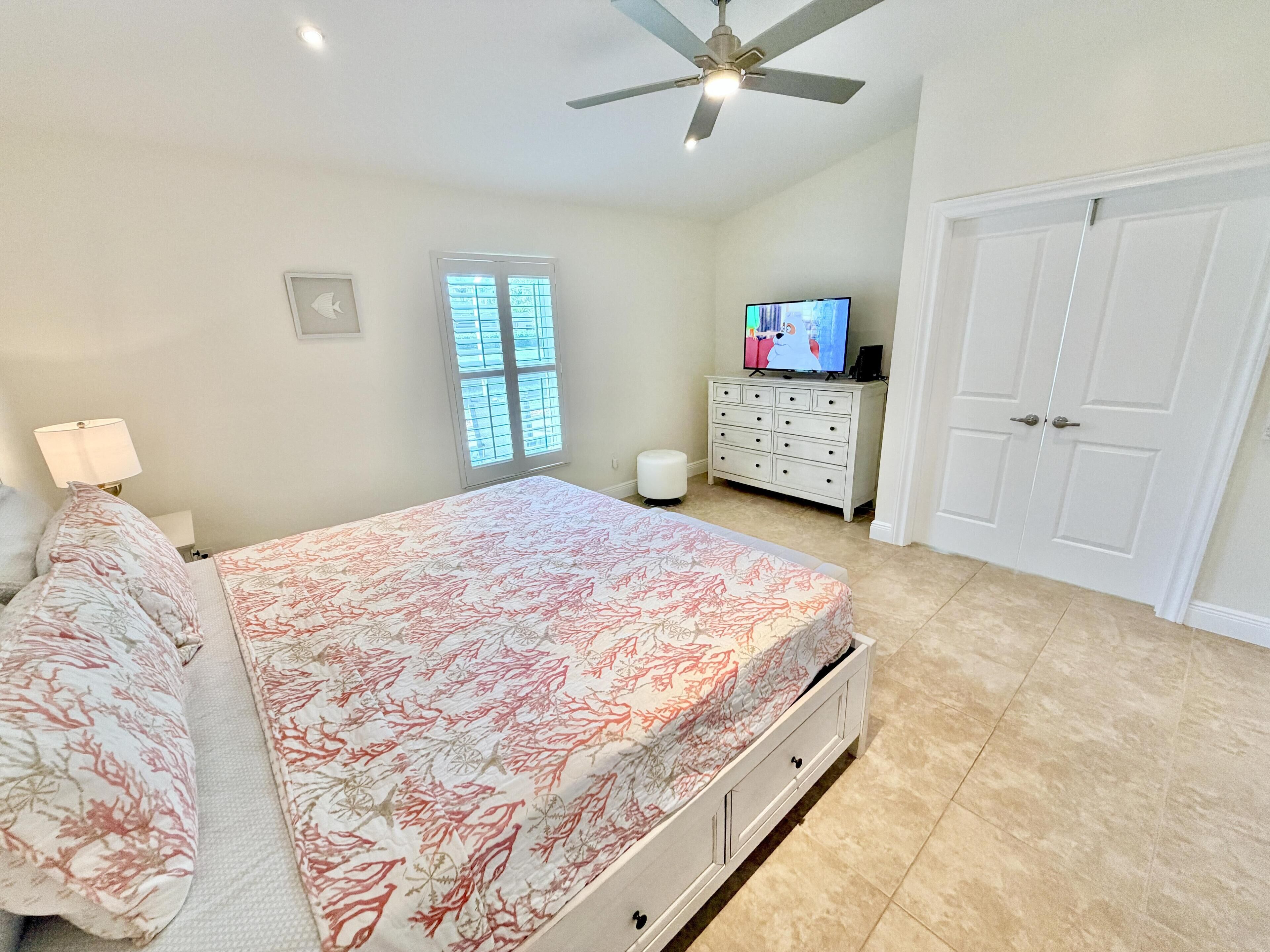 A bedroom with a bed, a ceiling fan, a dresser, a television, and a window.
