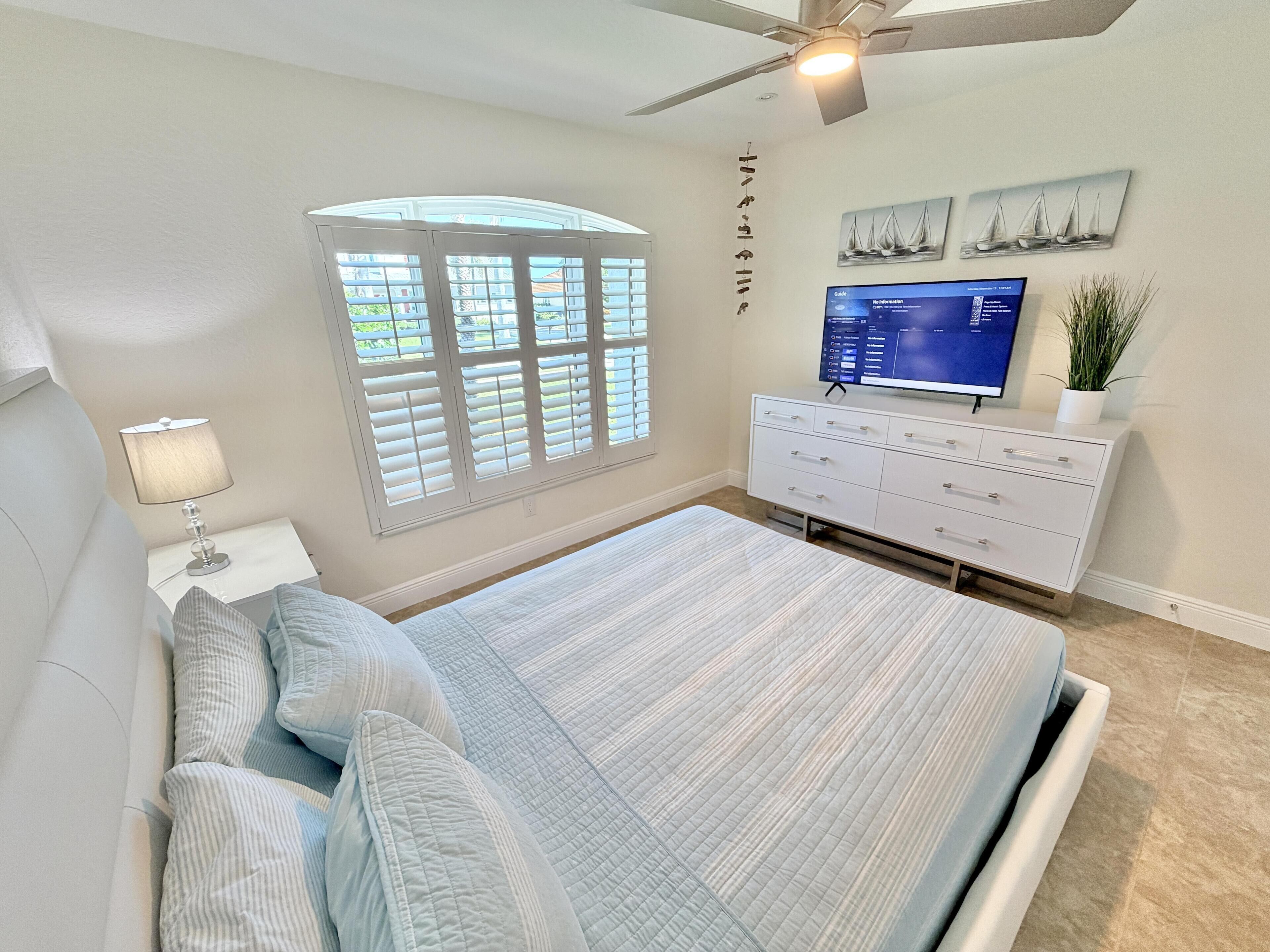 Un dormitorio con cama, mesita de noche, cómoda, televisor y ventana con persianas.