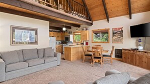 A cozy living room with a grey sofa, a wooden dining table with chairs, a kitchen area with wooden cabinets, and a large TV mounted on the wall. - Hansel Getaway Chalet – Cozy A-Frame Near Leavenworth! (Peshastin)
