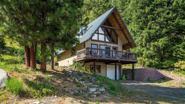 A two-story cabin with a balcony, surrounded by trees and a stone wall. - Hansel Getaway Chalet – Cozy A-Frame Near Leavenworth! (Peshastin)