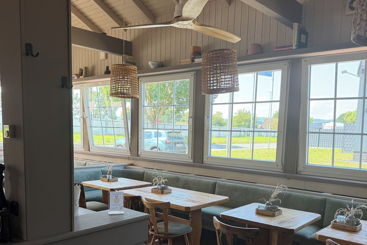 Un restaurant avec des tables et des bancs en bois, de grandes fenêtres donnant sur l’extérieur et des lampes en osier suspendues au plafond.