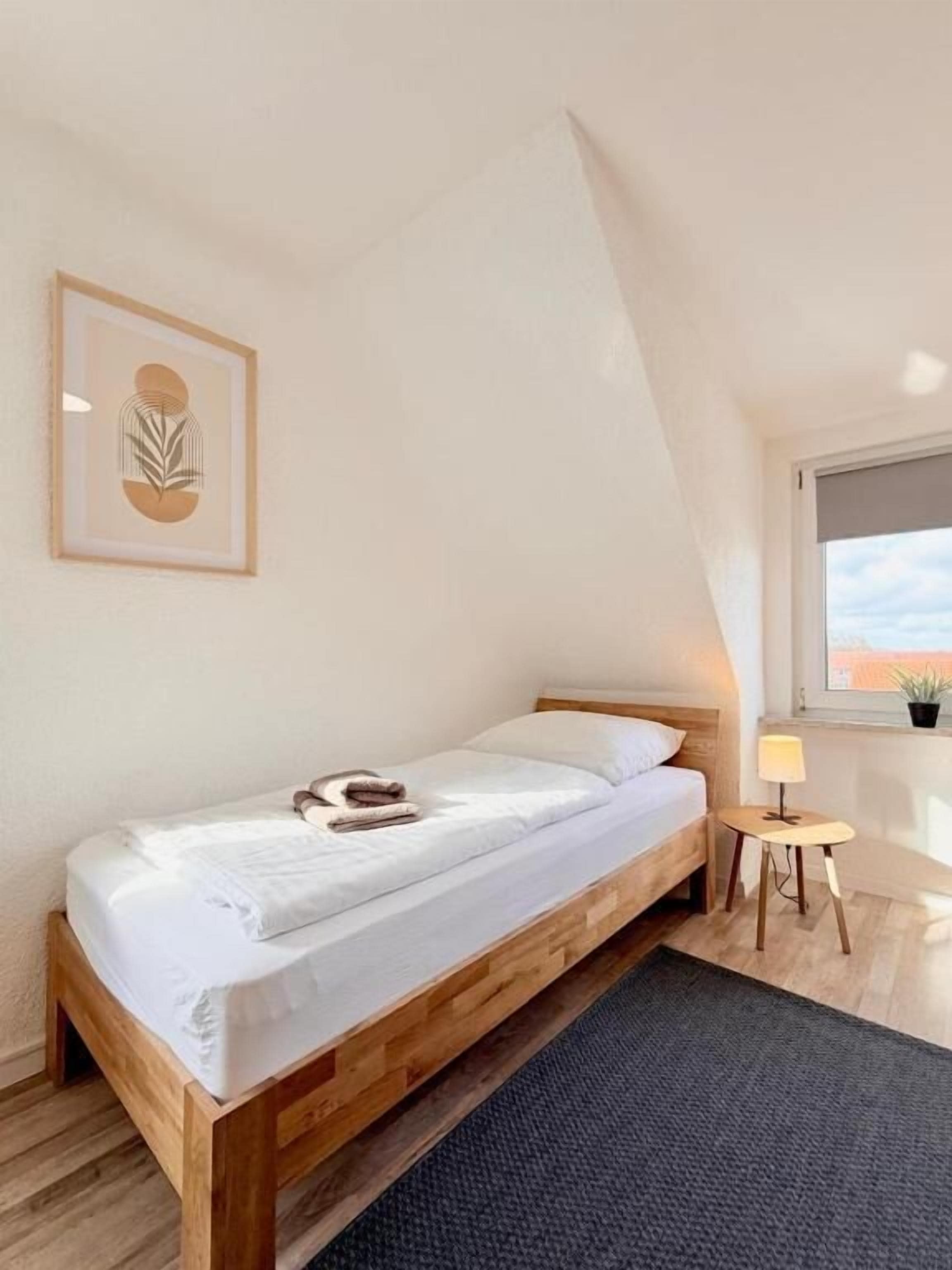 A single bed in a room with a sloped ceiling, a small table with a lamp, and a framed artwork on the wall.