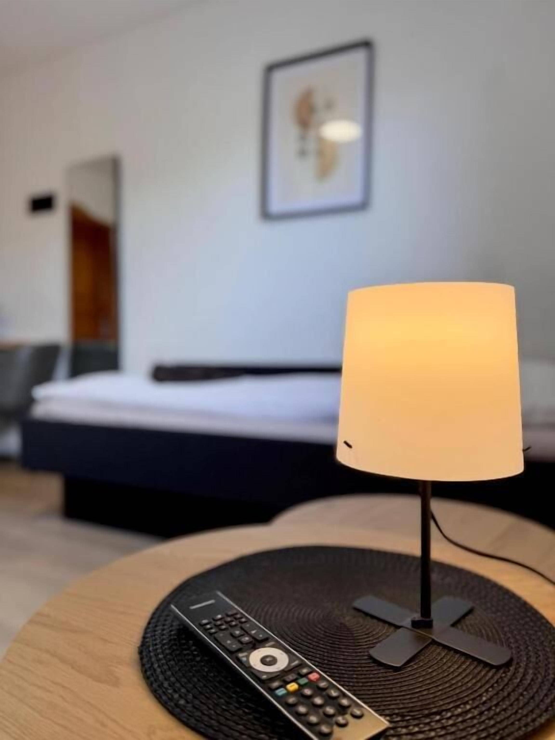 A hotel room with a bed, a bedside lamp, and a remote control on a round table.