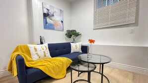 Een woonkamer met een blauwe bank, een gele plaid, een glazen salontafel en een potplant.