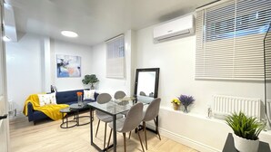 Een moderne woonkamer met een bank, salontafel en stoelen. Er is een eettafel met stoelen, een potplant en een lijst met spiegel.