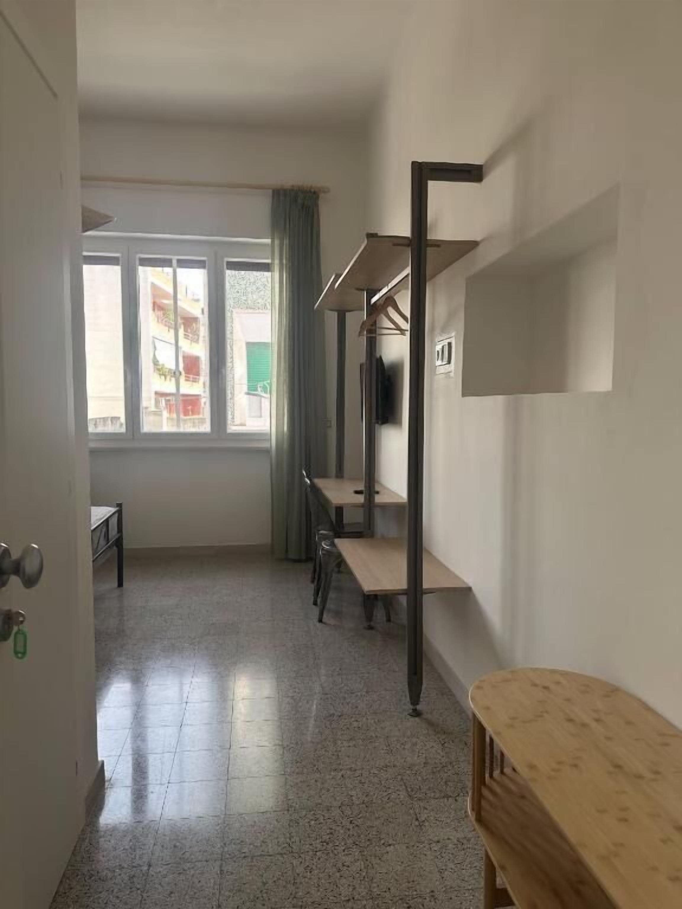 A room with a bed, a desk, and a chair. There is a window with curtains, and a door is visible on the left.