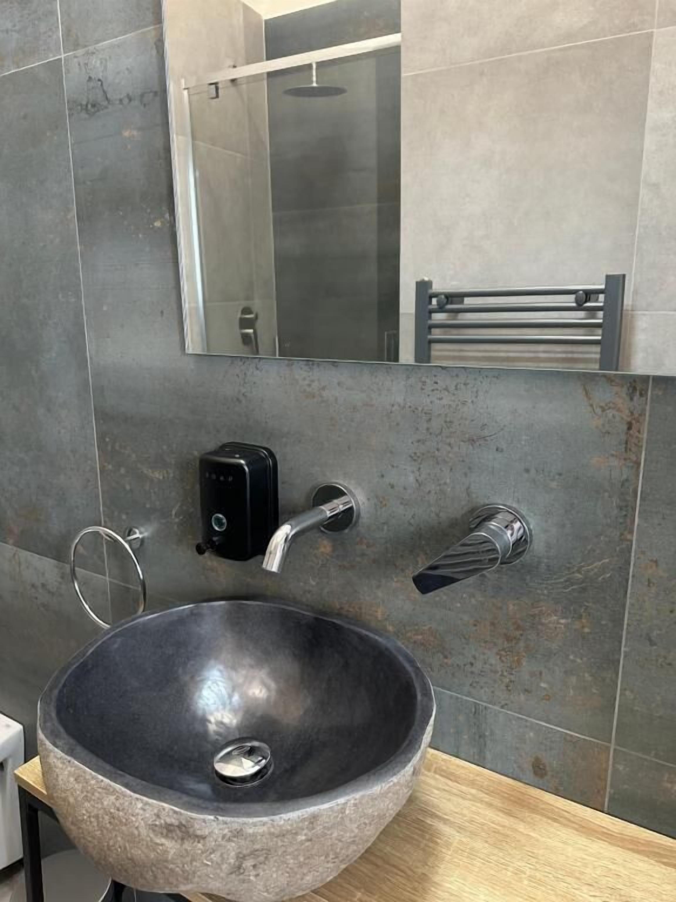 A modern bathroom with a large, round, stone sink, a handheld showerhead, a towel rack, and a mirror above the sink.