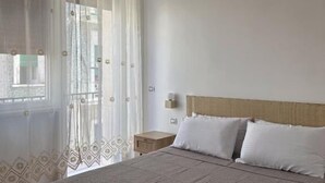 Une chambre à coucher avec un lit, une table de chevet, une lampe fixée au mur et des rideaux légers.