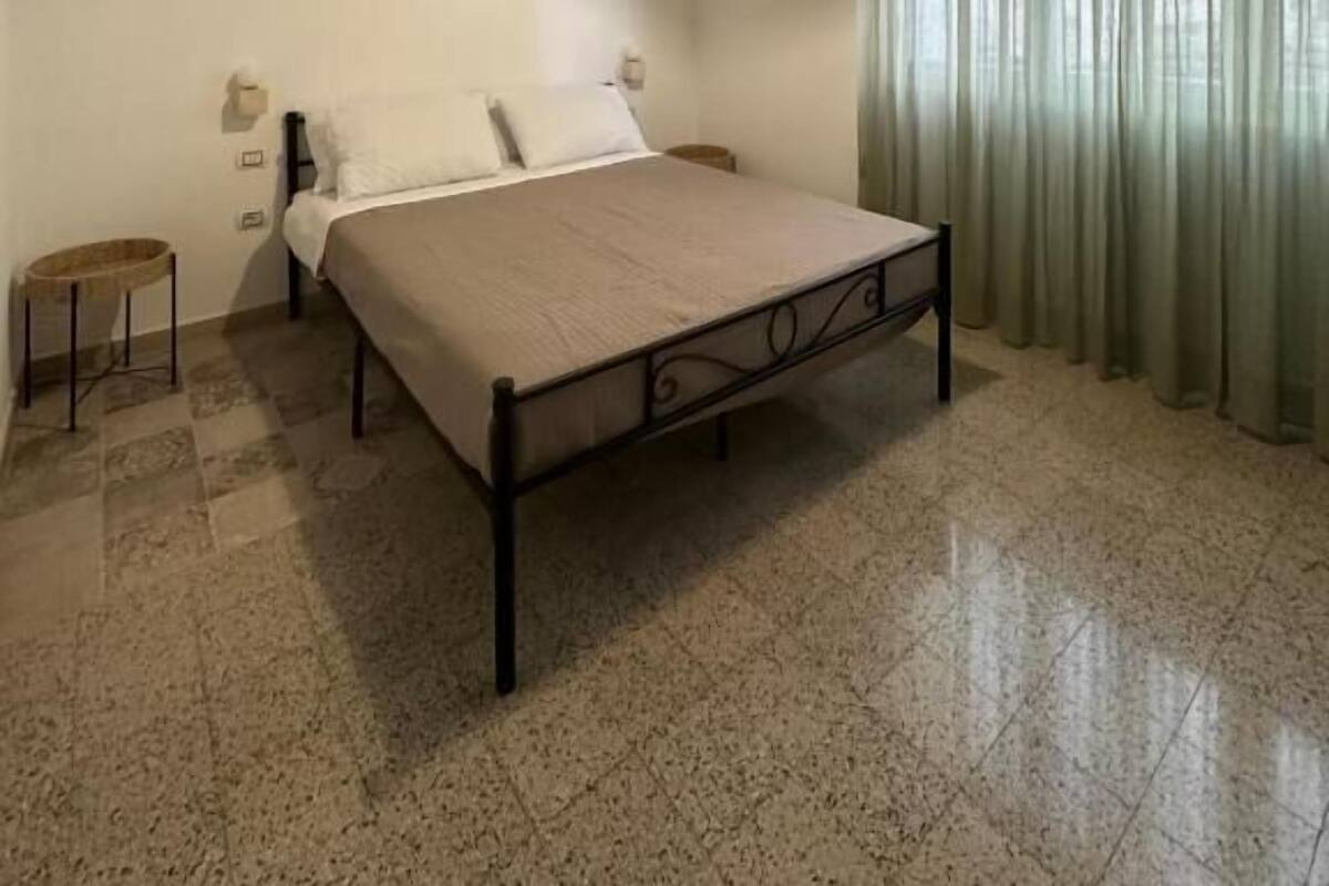 Ein Schlafzimmer mit einem Bett, einer Hängelampe, einem Fenster mit Vorhängen und einem kleinen Hocker.