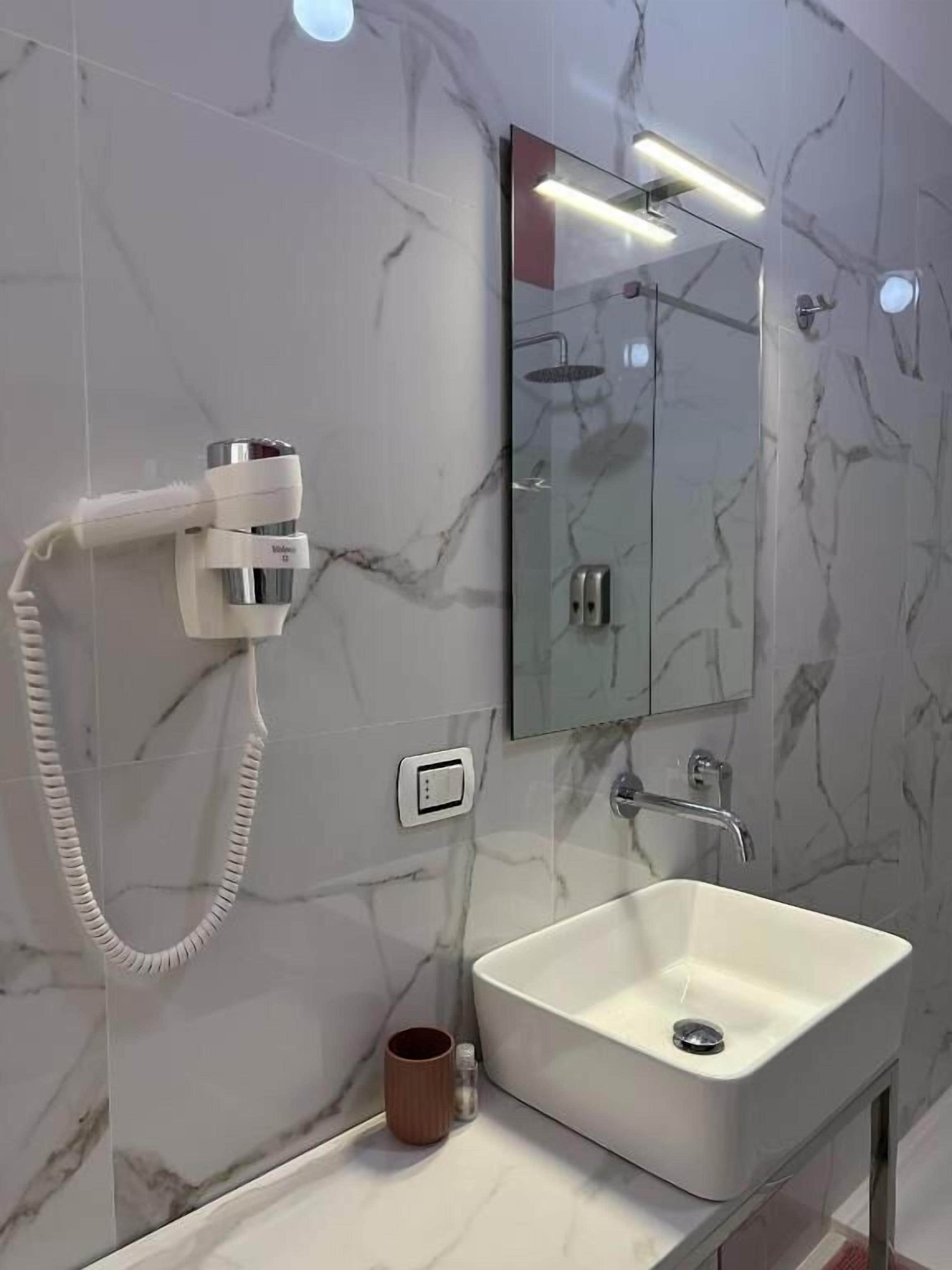 Un baño moderno con un lavabo blanco, un cabezal de ducha manual, un espejo y un dispensador de toallas.
