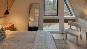 Une chambre avec un plafond en pente, un lit, une chaise, un miroir et une fenêtre donnant sur les arbres.
