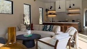 Amplia sala de estar con techo alto, vigas de madera, un conjunto de sofás, una mesa de centro y una zona de cocina con lámparas colgantes.