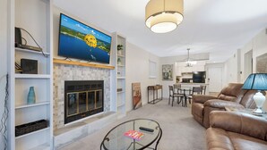 A living room with a fireplace, a television mounted on the wall, a dining area with a table and chairs, and a kitchen in the background. - Cozy Lakefront 2BR Condo Near HH, Golf, Dining & The Strip (Lake Ozark)