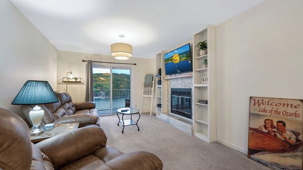 A modern living room with a sofa, a coffee table, a lamp, a fireplace, and a large window with a view of a lake. - Cozy Lakefront 2BR Condo Near HH, Golf, Dining & The Strip (Lake Ozark)