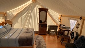 A cozy tented accommodation with a bed, a wooden dresser, a small table, a chair, and a fan.