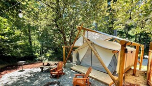 A wooden tent with a canvas roof, surrounded by gravel and trees. There are two wooden chairs and a small table nearby.