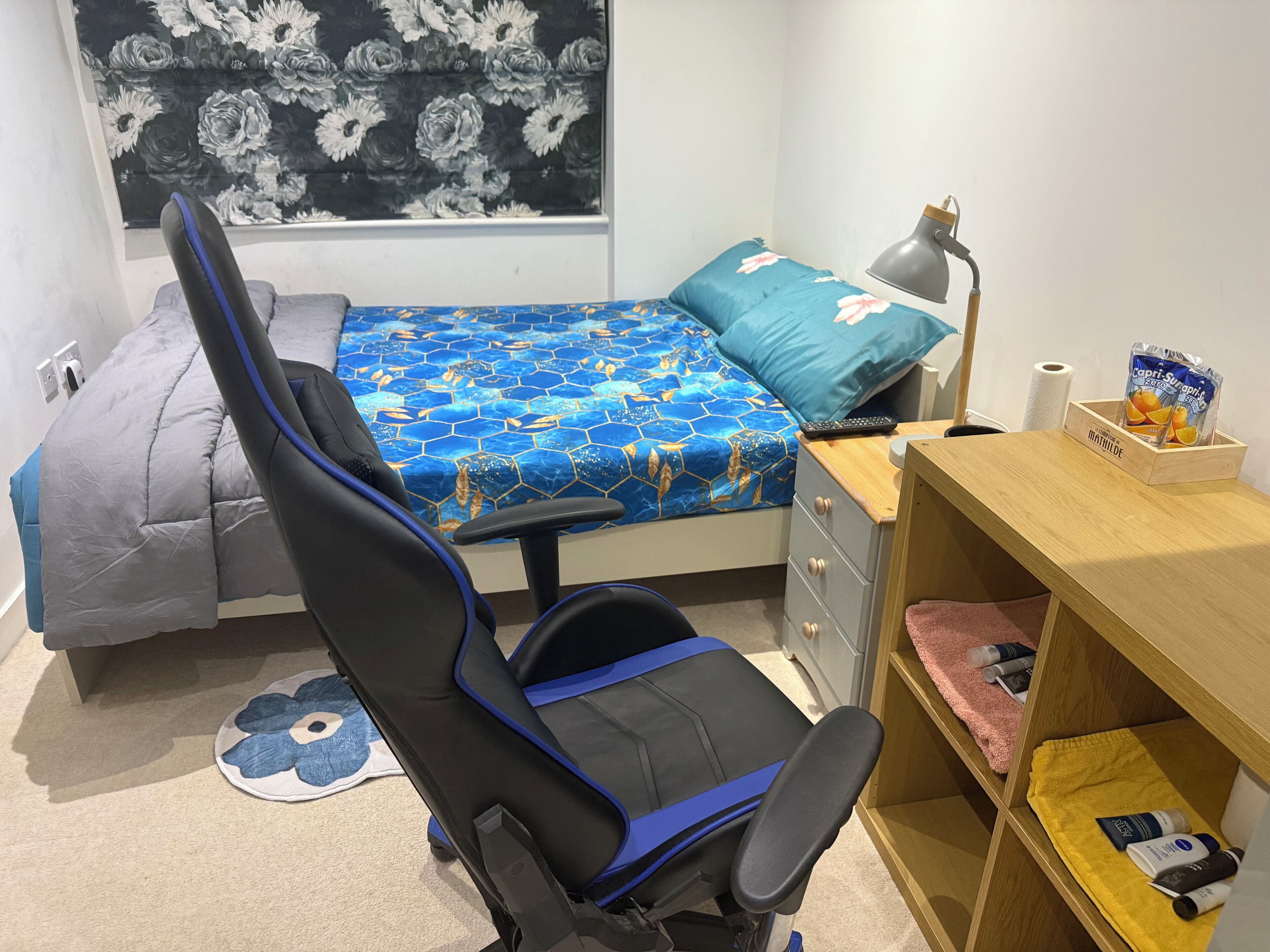 A bedroom with a bed, a desk, a chair, a lamp, and a shelf with toiletries.