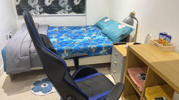 A bedroom with a bed, a desk, a chair, a lamp, and a shelf with toiletries.