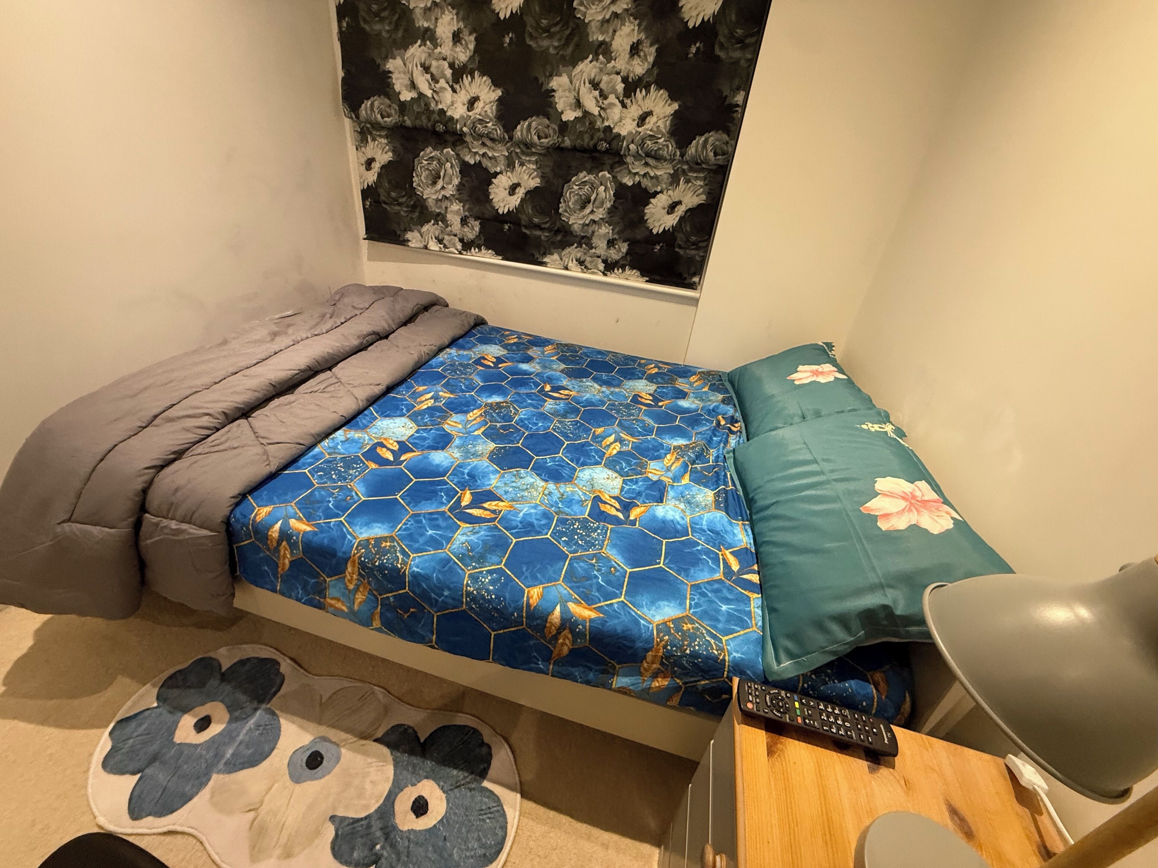 Quarto pequeno com cama, janela com cortinas florais, luminária de cabeceira e controle remoto sobre uma mesinha de madeira.