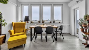 Un comedor con una mesa de madera dispuesta para cuatro personas, rodeada por cuatro sillas. Hay ventanas grandes con marcos blancos, un radiador debajo de la mesa y plantas en macetas en el alféizar.
