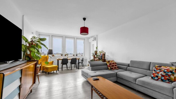 Una sala de estar moderna con un sofá gris, una mesa de centro de madera y un televisor sobre un mueble de madera. Hay sillones amarillos, una mesa de comedor y ventanas amplias con vistas.