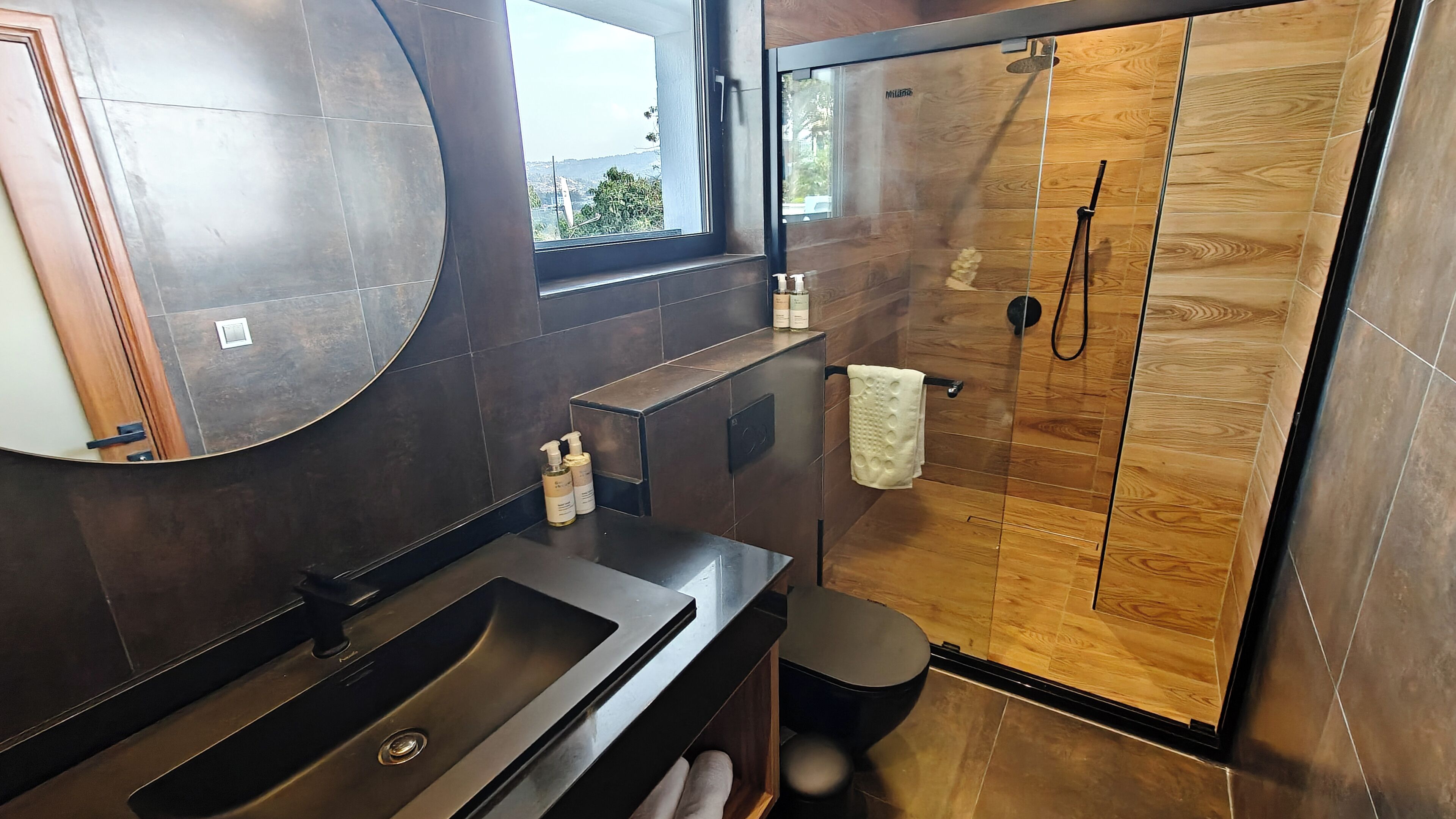 A modern bathroom with a large mirror, dark tiles, a wooden shower enclosure, and a window with a view.