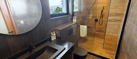 A modern bathroom with a large mirror, dark tiles, a wooden shower enclosure, and a window with a view.