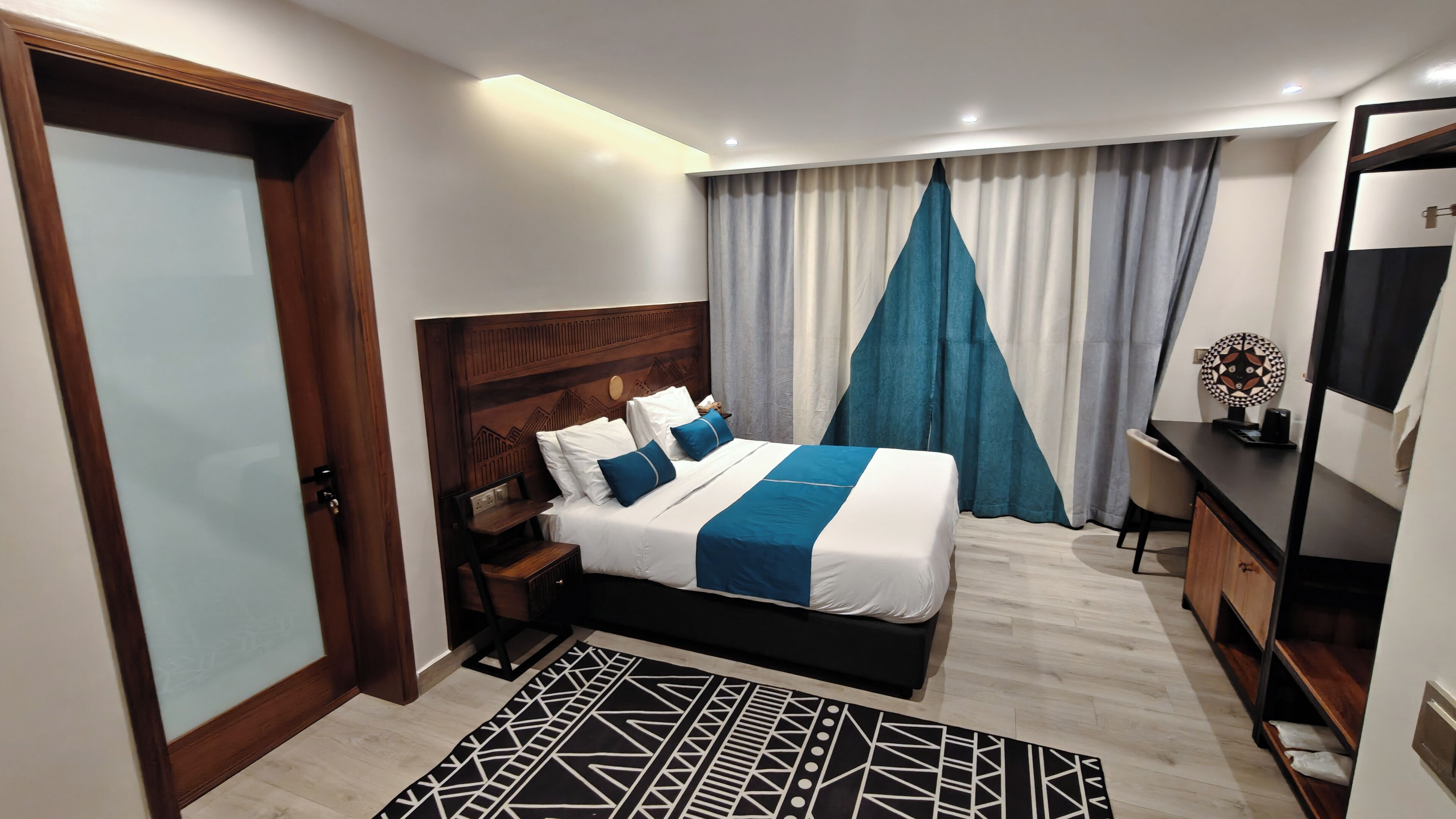 A modern hotel room with a large bed, a wooden headboard, a desk, a chair, a TV, and a window with curtains.