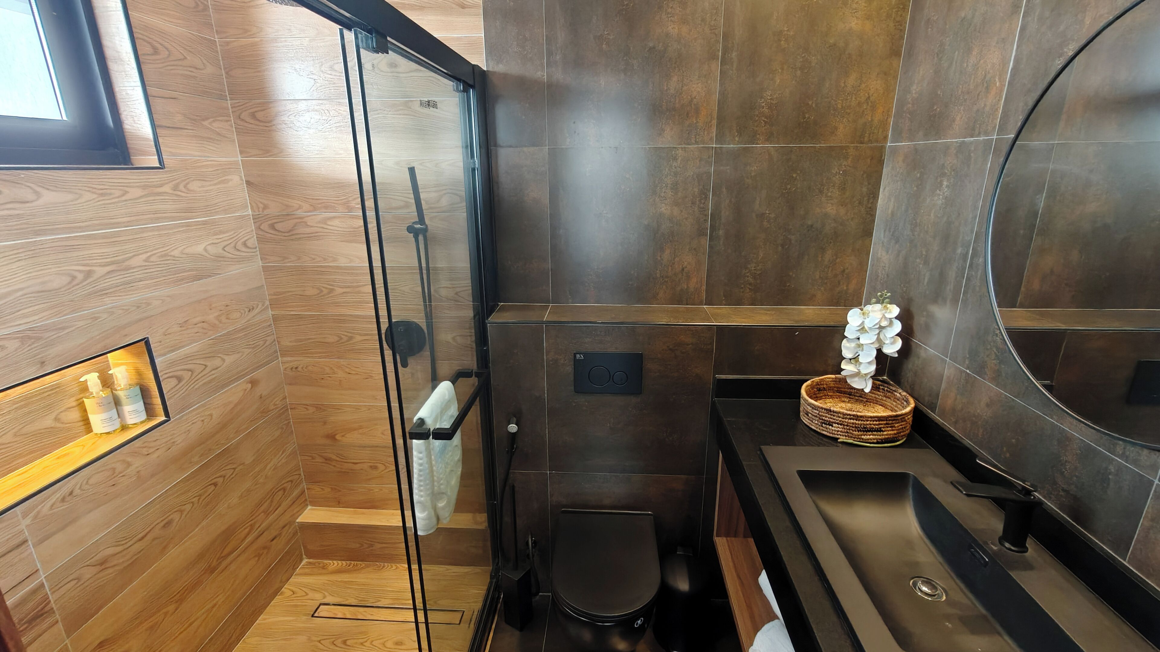 A modern bathroom with a wooden floor, a glass shower enclosure, a black toilet, and a sink with a woven basket and white flowers on the counter.