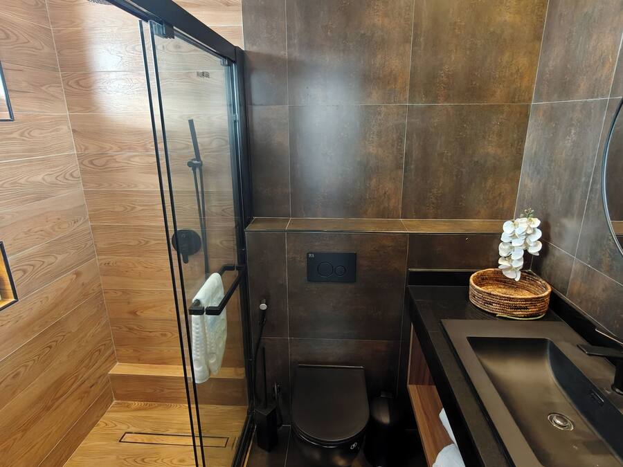 A modern bathroom with a wooden floor, a glass shower enclosure, a black toilet, and a sink with a woven basket and white flowers on the counter.