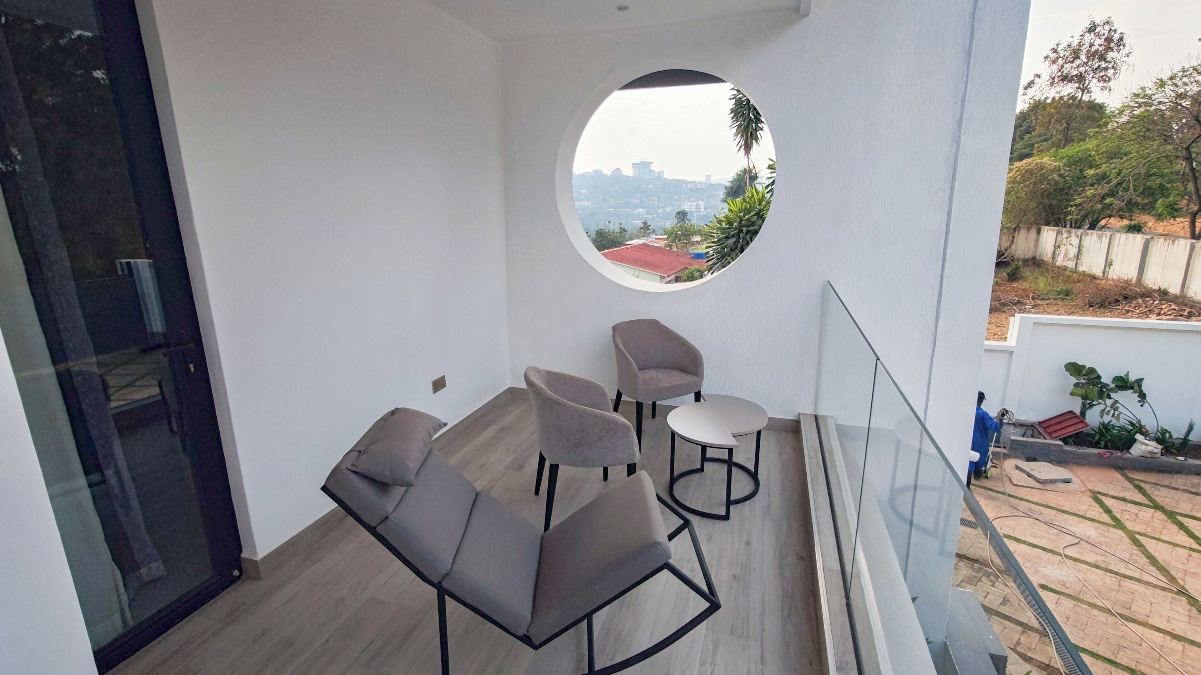 A balcony with a round window, two grey chairs, a small table, and a view of a cityscape.