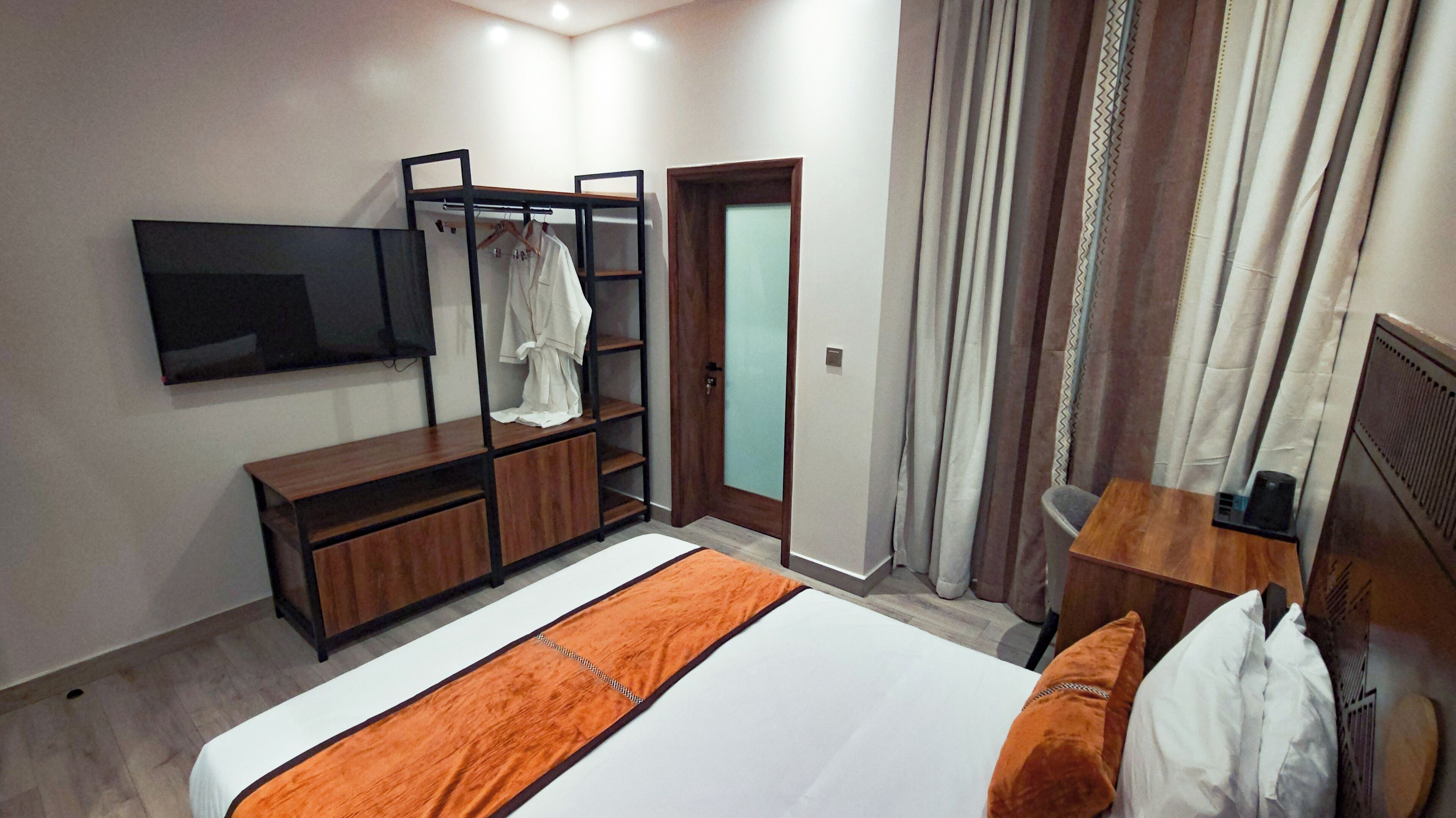 A hotel room with a bed, a TV mounted on the wall, a wooden dresser, a robe rack, and a door with curtains.