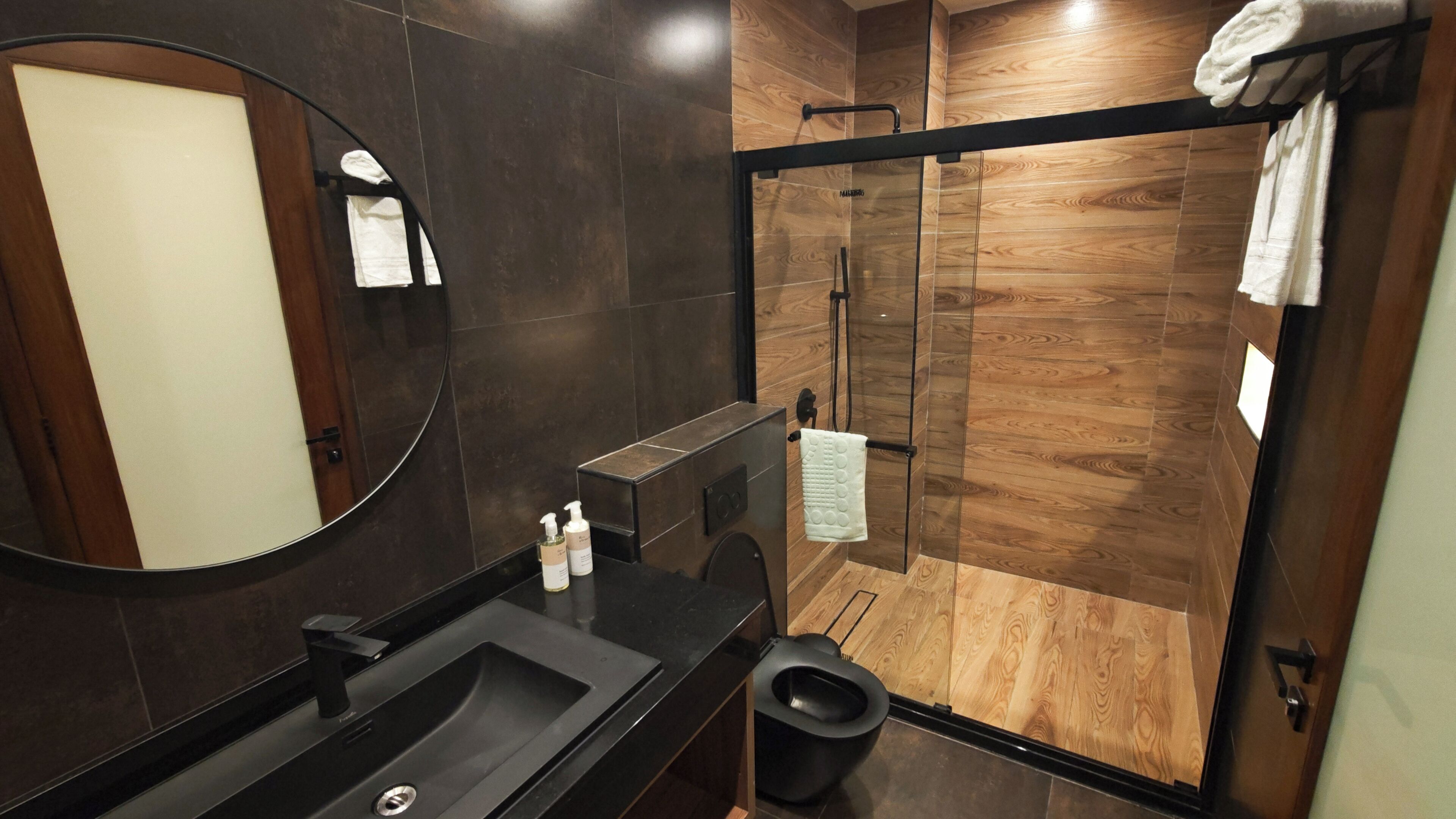 Une salle de bain moderne avec un grand miroir, des carreaux foncés, un lavabo noir et une cabine de douche en verre.
