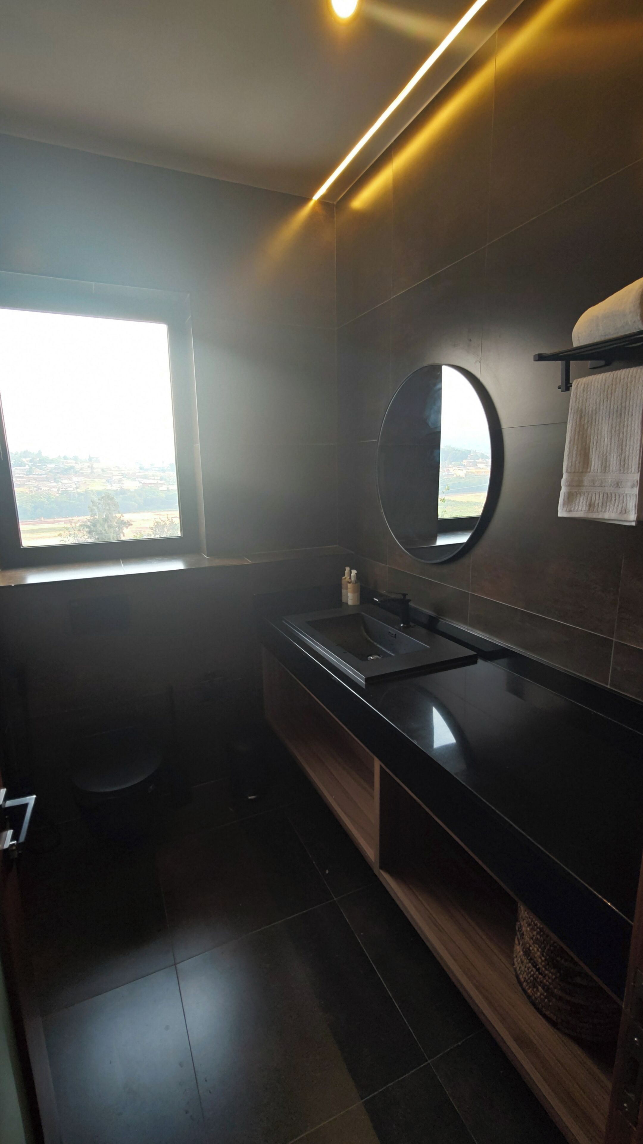 A modern bathroom with a large window, a round mirror, and a black countertop.