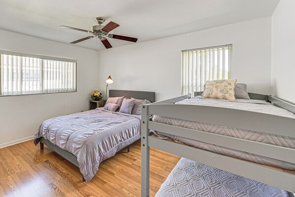 Une chambre avec un lit superposé, un ventilateur de plafond, deux fenêtres équipées de stores, une table de chevet avec une lampe et une petite table de nuit avec un vase de fleurs.