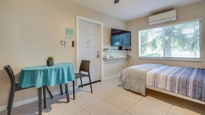 Una habitación pequeña y bien iluminada con una cama, una mesa con una planta, una silla, un ventilador de techo y una ventana con persianas.