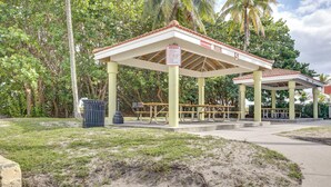 Un área de picnic cubierta con bancos y un contenedor de basura, rodeada de follaje.