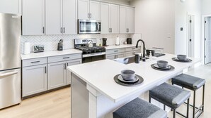 Een moderne keuken met witte kasten, een roestvrijstalen koelkast, een magnetron en een gasfornuis. Er staan drie barkrukken bij het eiland.