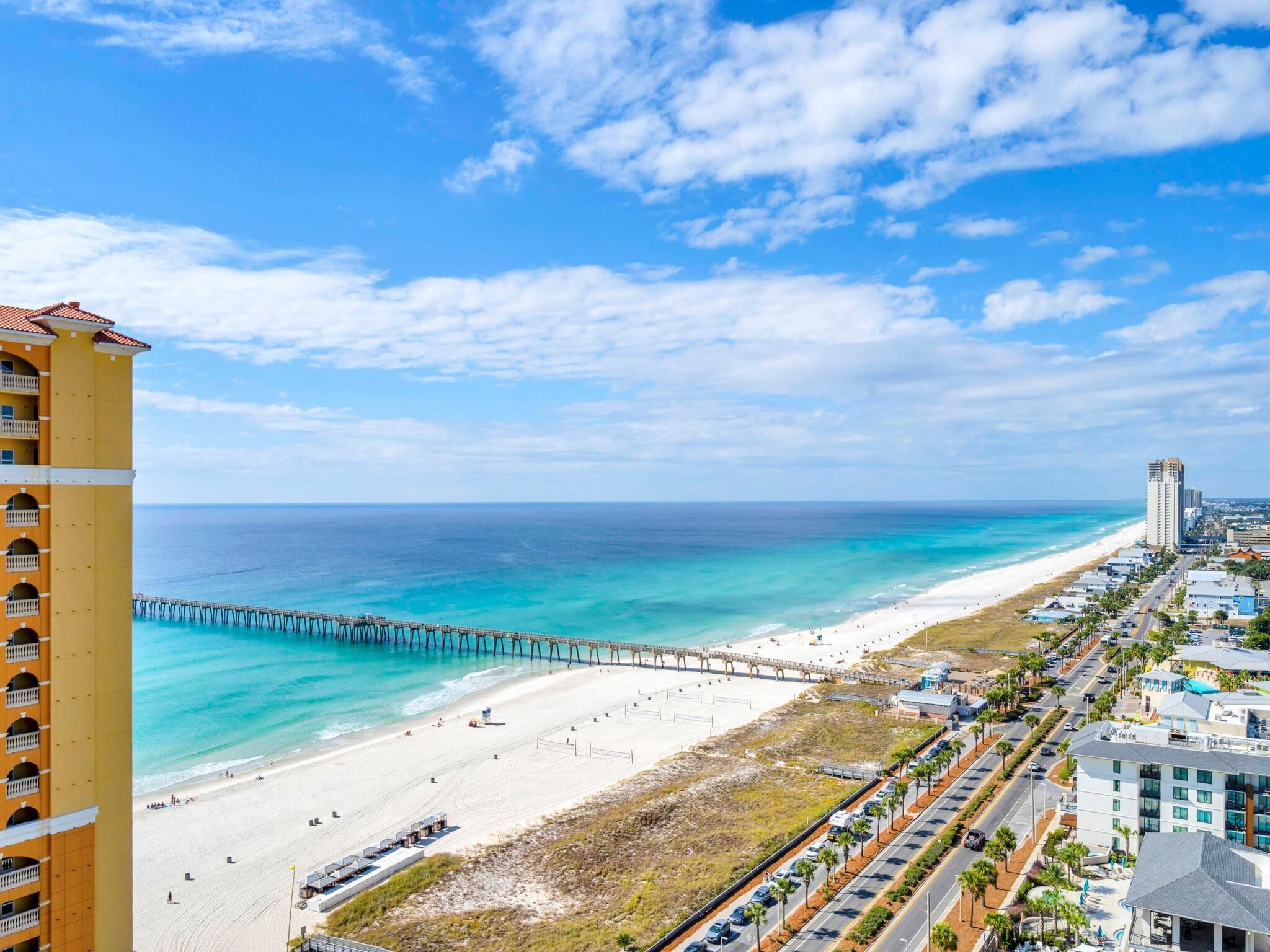 A coastal hotel with a beach view, a pier, and a road lined with palm trees.