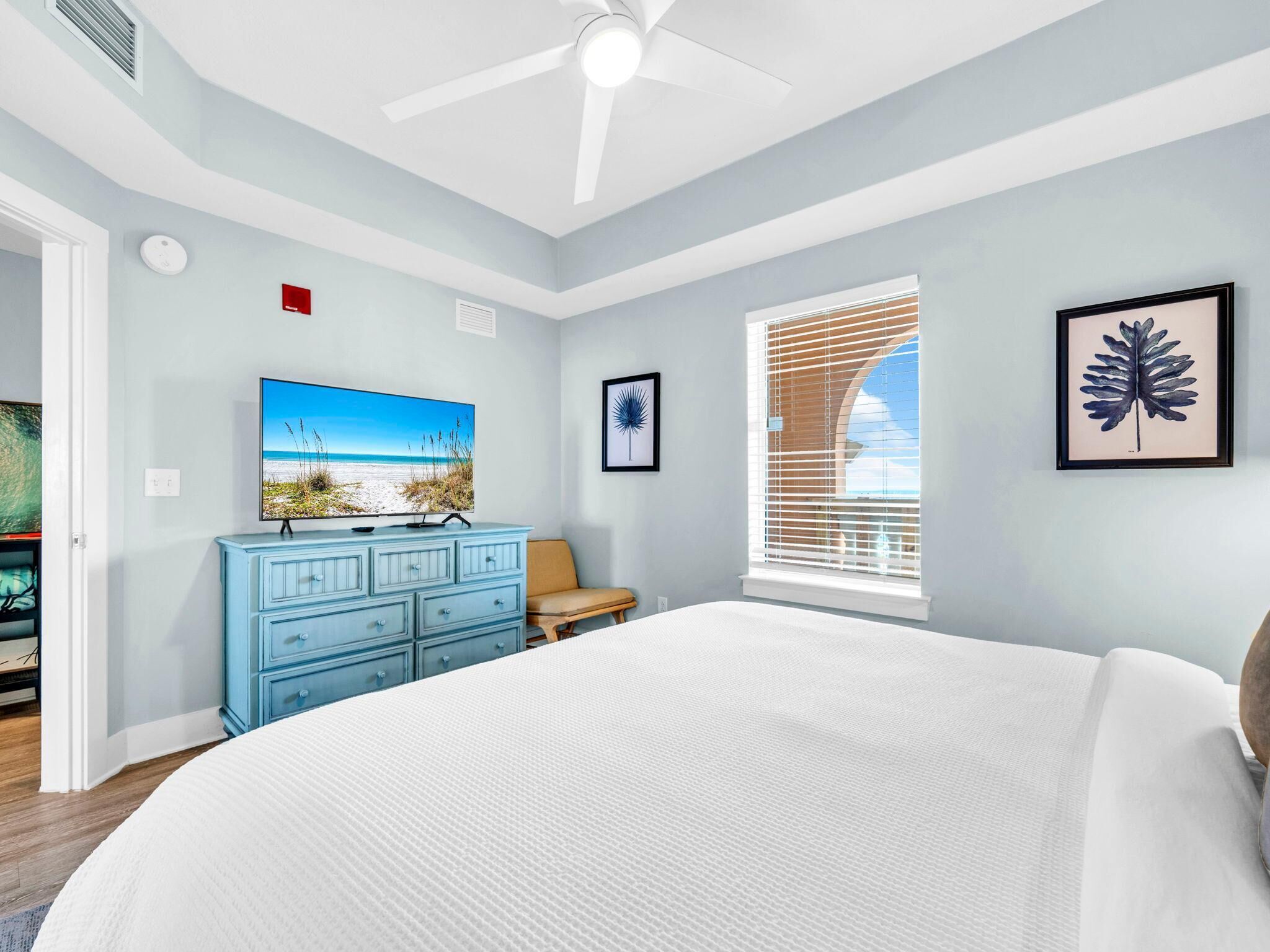 Een slaapkamer met een bed, een blauwe dressoir, een televisie waarop een strandafbeelding te zien is, een raam met jaloezieën en een plafondventilator.