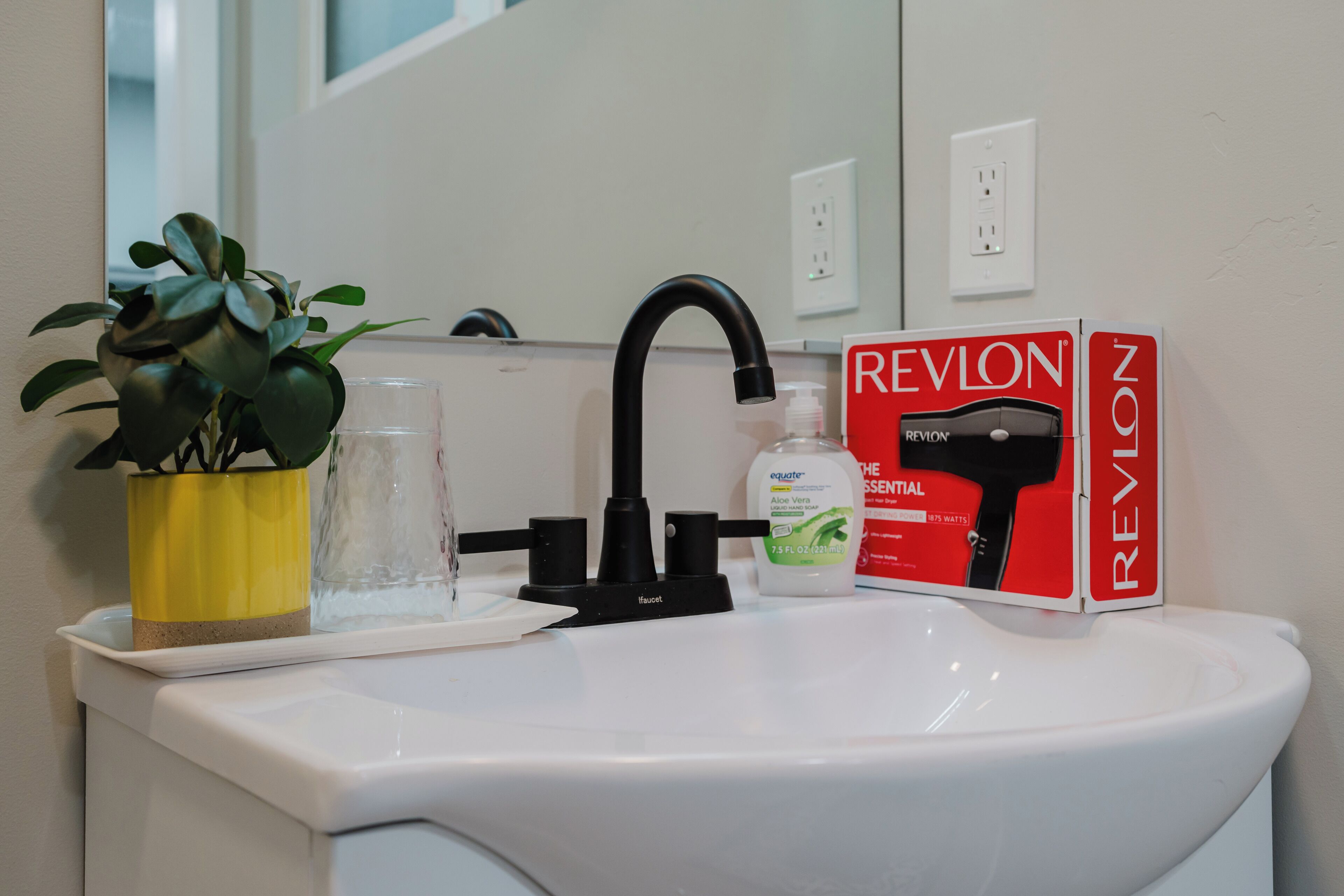 Ein Waschbecken mit schwarzem Wasserhahn, ein Blumentopf, ein Glas, eine Handseifenflasche und eine Box für einen Revlon-Haartrockner.