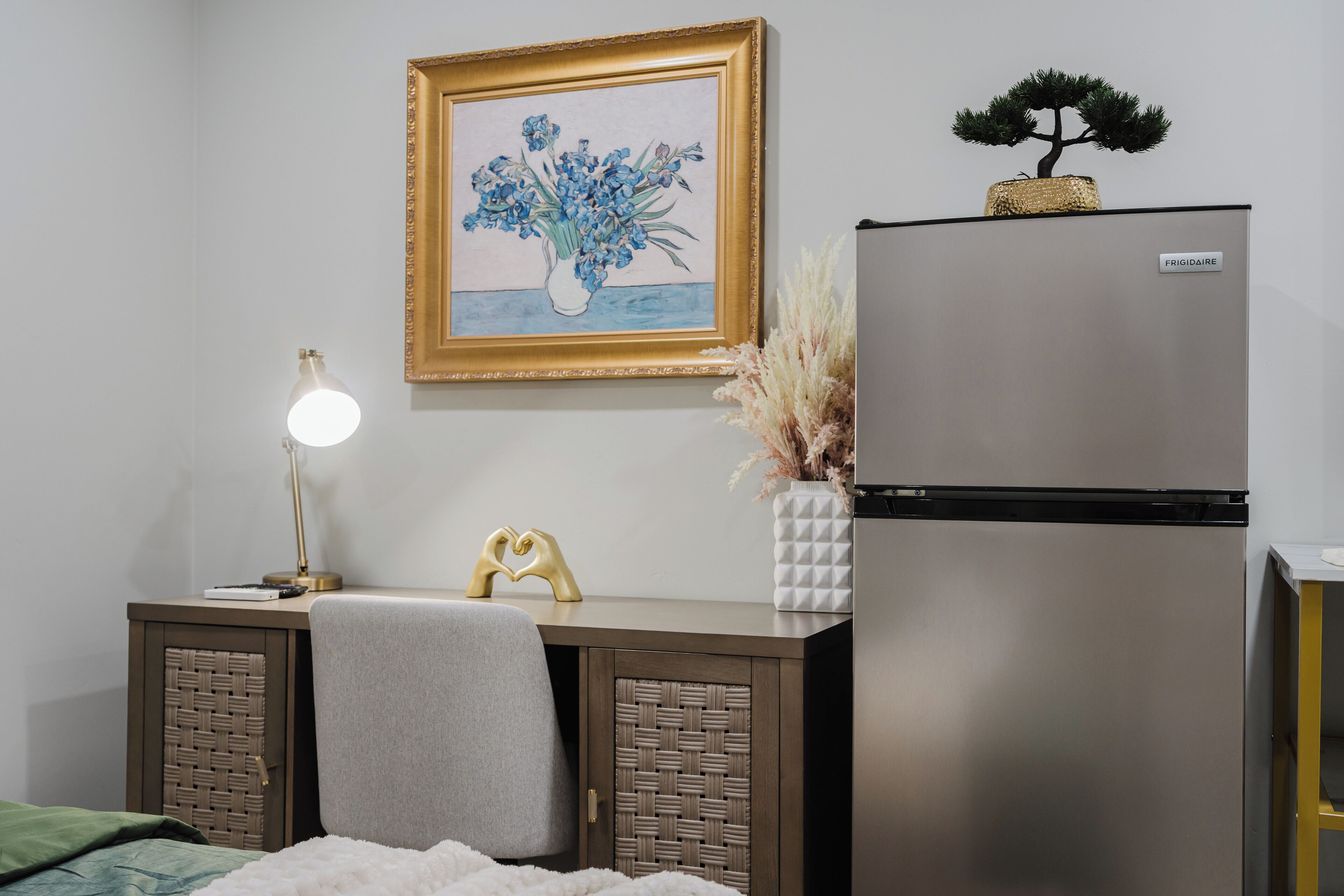 A bedroom with a bed, bedside table, lamp, framed artwork, and a refrigerator.