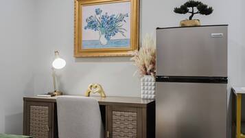 A bedroom with a bed, bedside table, lamp, framed artwork, and a refrigerator.