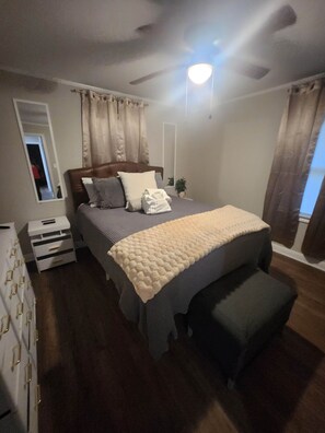 A bedroom with a bed, a ceiling fan, a nightstand, a dresser, and a window with curtains. - FrankleeB Haven Andrews (Andrews)