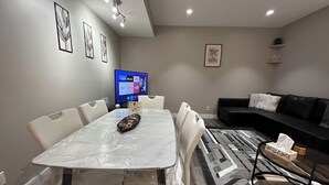 Una sala de estar moderna con una mesa de comedor, sillas blancas, un sofá negro y un televisor de pantalla plana montado en la pared.