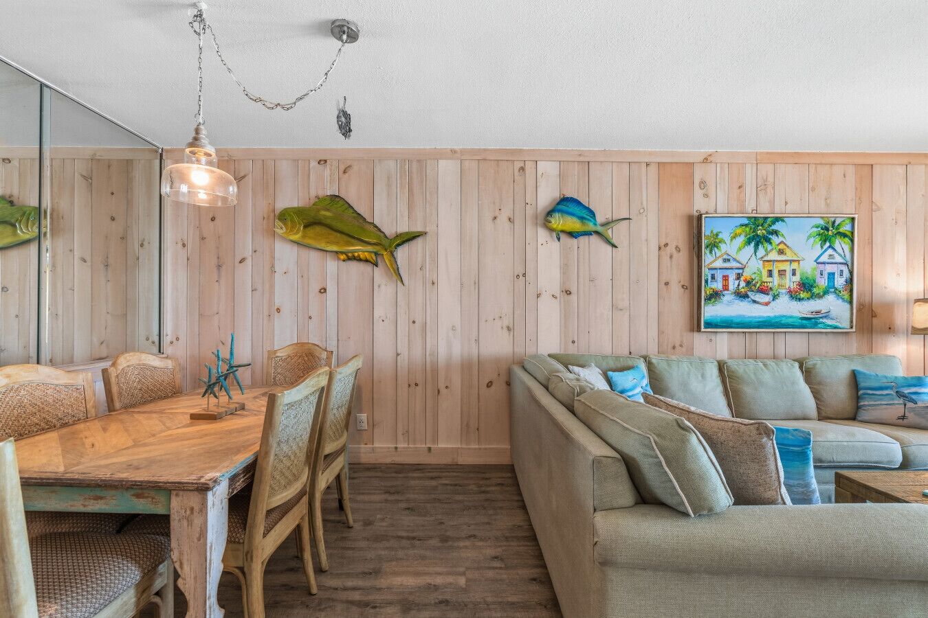 Une pièce aux murs recouverts de lambris en bois, une table à manger avec des chaises, un canapé et un tableau représentant une scène de plage.