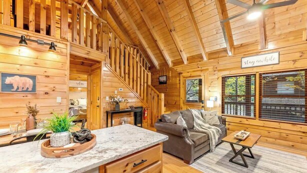 A cozy log cabin interior with a wooden staircase, a dining area, and a living room with a couch and a coffee table. - Hidden Cozy Cabin Villa with Hot Tub Close to Everything (Pigeon Forge)