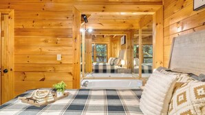 A cozy wooden cabin interior with a checkered floor, a bed with a plaid blanket, a small table with books and a plant, and a view of a deck and outdoor area. - Hidden Cozy Cabin Villa with Hot Tub Close to Everything (Pigeon Forge)