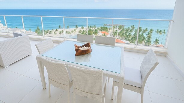 A balcony with a glass table and wicker chairs, overlooking a beach with palm trees and the ocean. - Hemingway By YellowKey (Guayacanes)